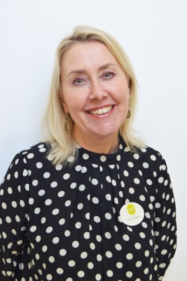 Portrait of Sarah Rogers, smiling with blonde hair, wearing a black polka dot blouse and a white floral brooch, standing against a plain white background.