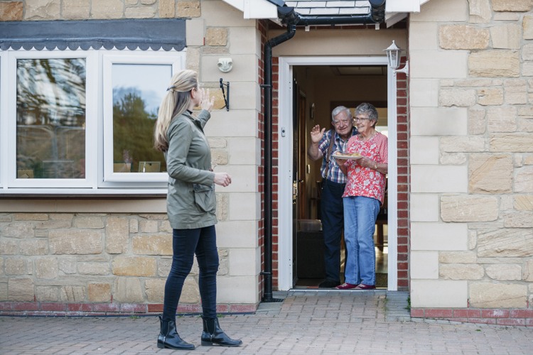 A woman stands outside a home, waving to the elderly couple standing in the open doorway
