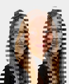 A person with long, straight, light brown hair standing against a plain light background. They are wearing a black top and a round badge on their chest