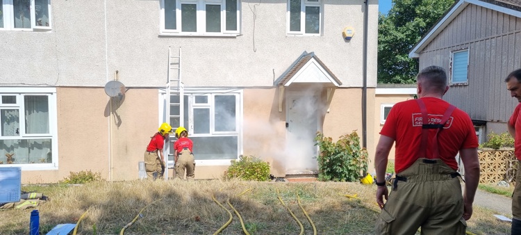 Smoke escapes from a house as firefighters and team members monitor the situation.