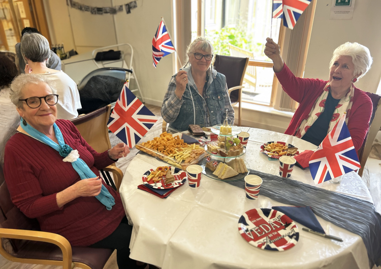 Three elderly ladies, waving patriotic Union Jack flags, smiling and laughing together as they sit around a small round table, enjoying an afternoon tea.