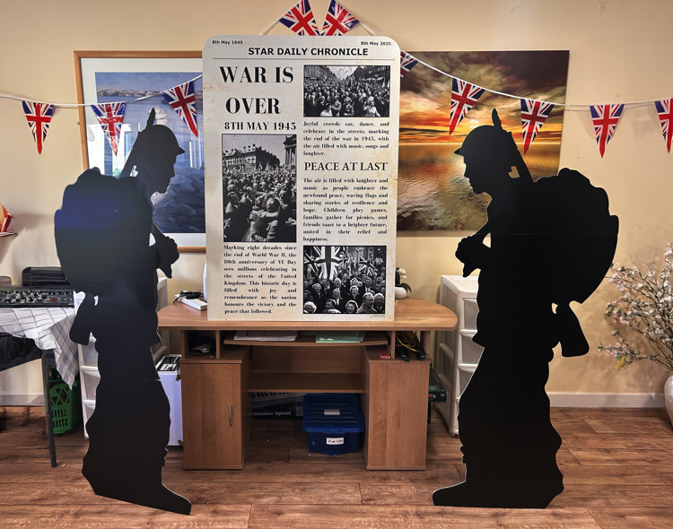 A large VE Day display features a mock newspaper front page with the headline “WAR IS OVER – 8th May 1945”, flanked by two life-size black silhouettes of World War II soldiers. The setup is indoors, decorated with Union Jack bunting, and includes a sunset painting and a large photo of wartime celebrations in the background. The display commemorates the 80th anniversary of VE Day.