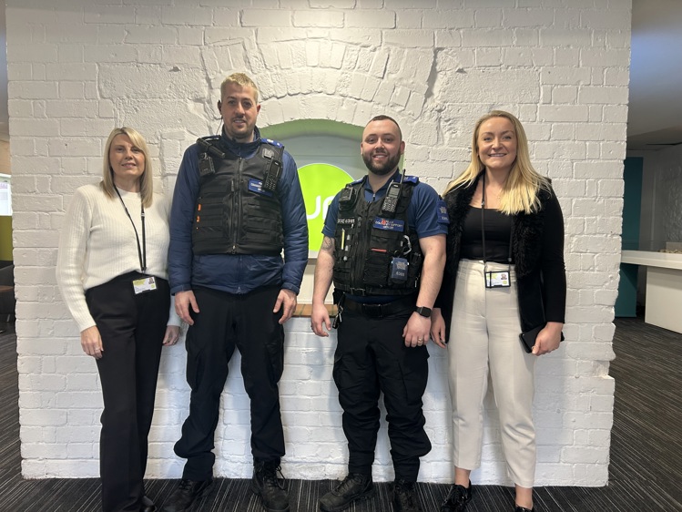 A group of four people stands in front of a white brick wall with a green Curo logo in the background. Two individuals in the center are wearing black police-style vests with radios and ID badges, while the two women on either side are dressed in business casual attire with lanyards. All are smiling and looking at the camera. The setting appears to be an indoor office or community space.