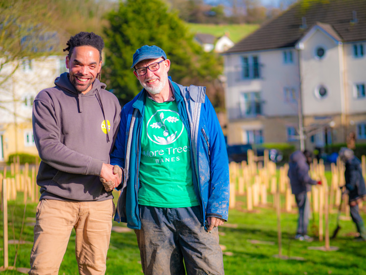 Two men shake hands in front of some newly planted trees and shrubs