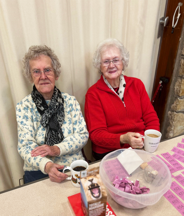 Two elderly ladies, both with short grey curly hair and glasses, one in a red cardigan and the other in a floral top, sitting drinking tea.