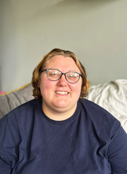 Abs Clements smiling wearing glasses, with short dark blonde hair wearing a navy tshirt.