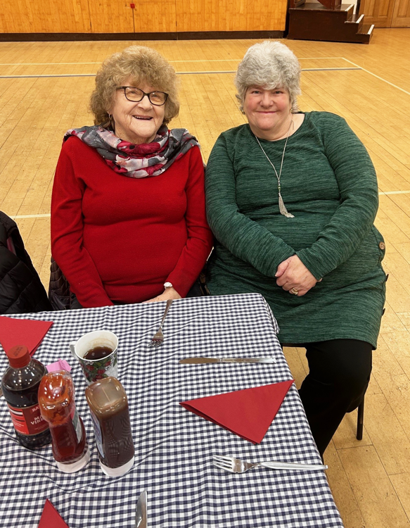 Two smiling elderly ladies, one with short curly brown hair and a red jumper, the other with short grey hair and a green jumper, sitting at a table