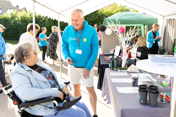 A person in a wheelchair speaks with a Curo employee wearing a turquoise hoodie and lanyard beside a table displaying event materials. The scene is set under a marquee at an outdoor community event, with additional attendees, decorations and display stands visible in the background.