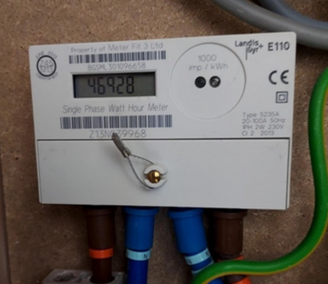 Digital electricity meter mounted on a wall, showing an electricity usage reading on an electronic display, with coloured cables connected below.